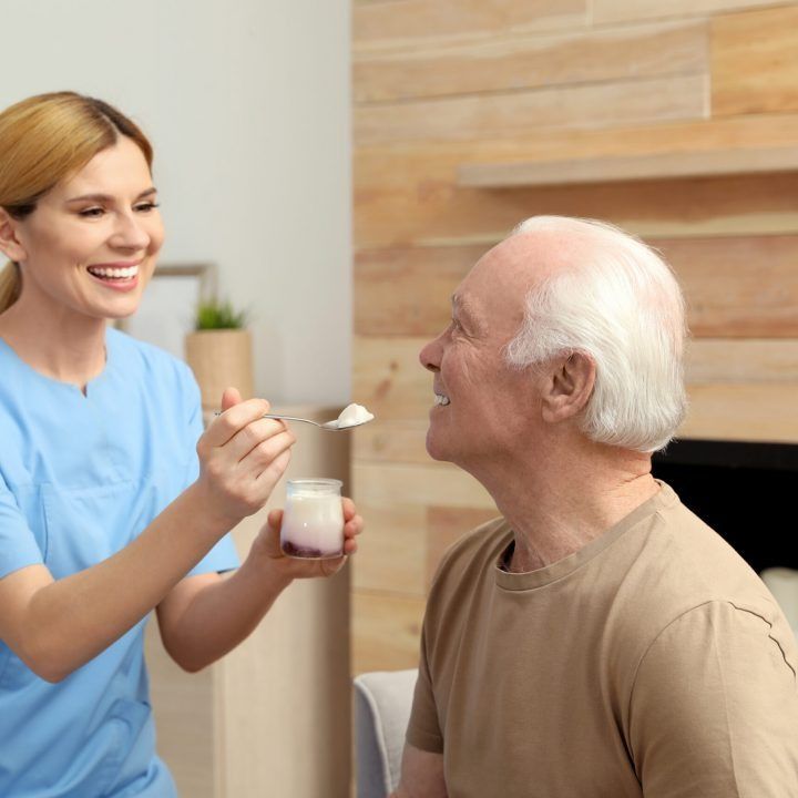 Caregiver offers a spoonful of yogurt to a smiling resident in a bright care home room