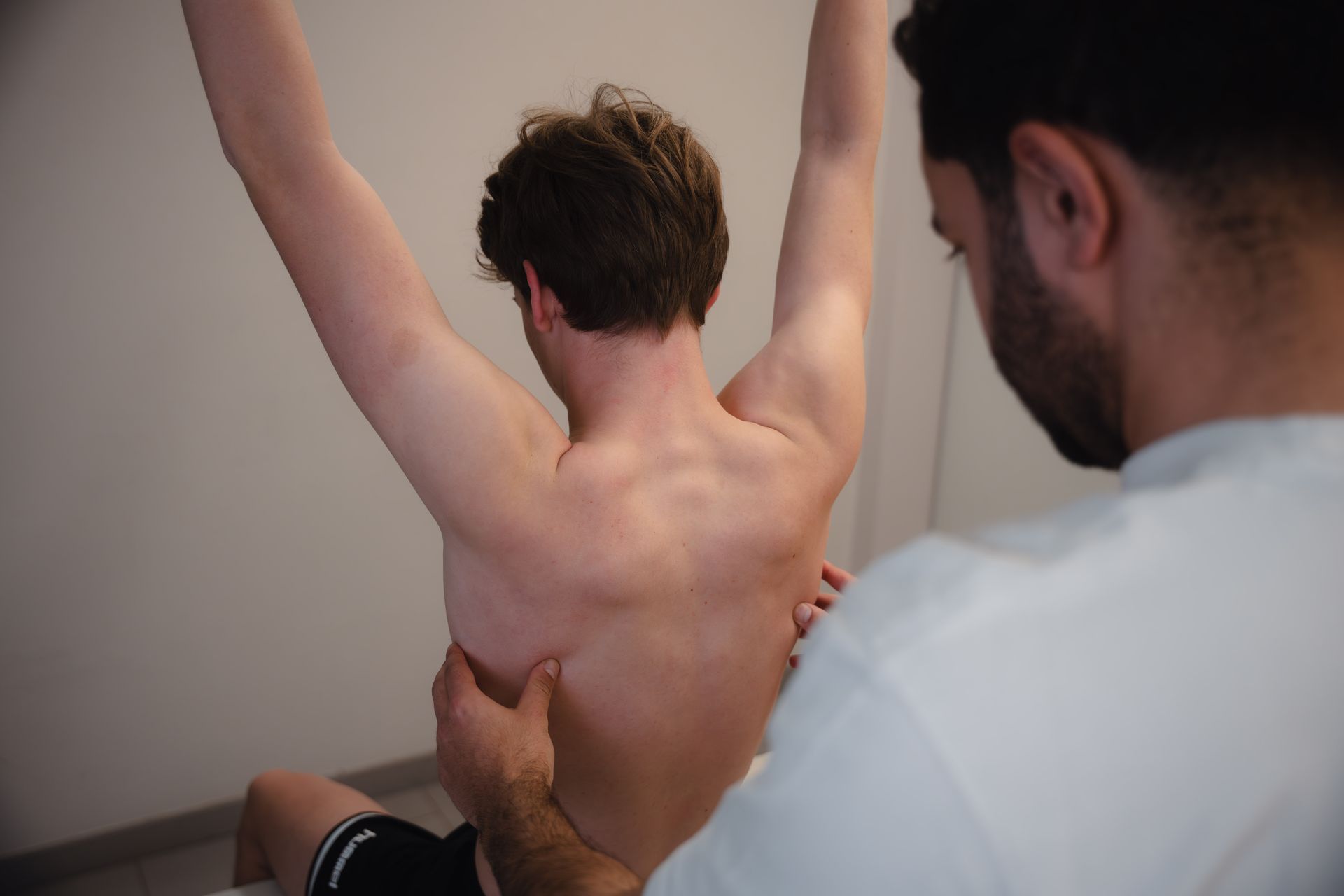 Een persoon zit met opgeheven armen en ondergaat in een witte kamer een rugonderzoek door een zorgverlener.