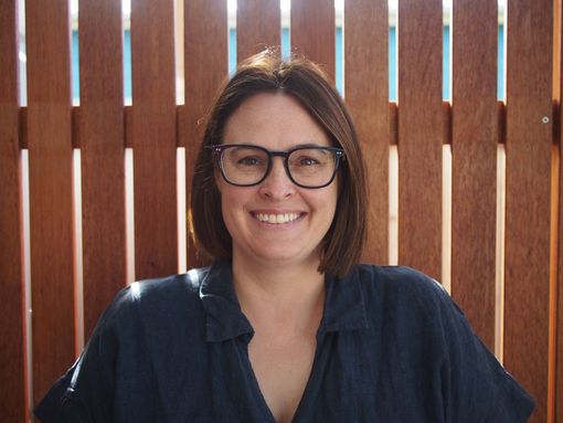 Smiling person with glasses in a navy shirt, standing before a wooden fence