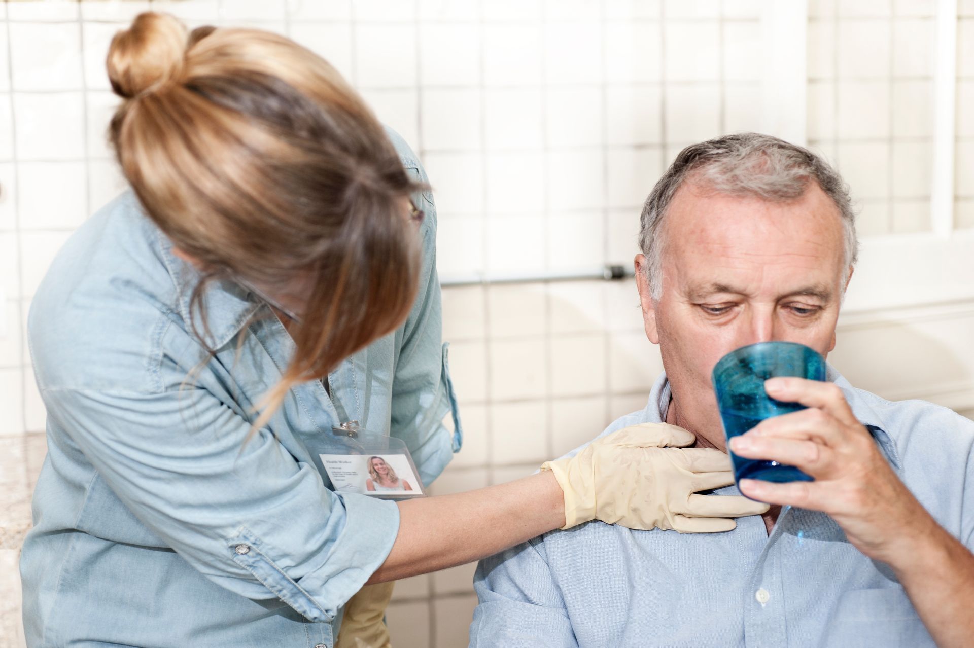 Caregiver gently cleans an older man’s face with a cloth while he drinks from a blue cup.