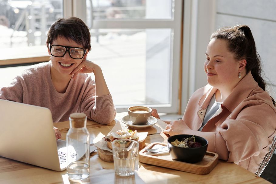 Two people chatting at a café table with laptops, coffee, and breakfast dishes by a bright window.