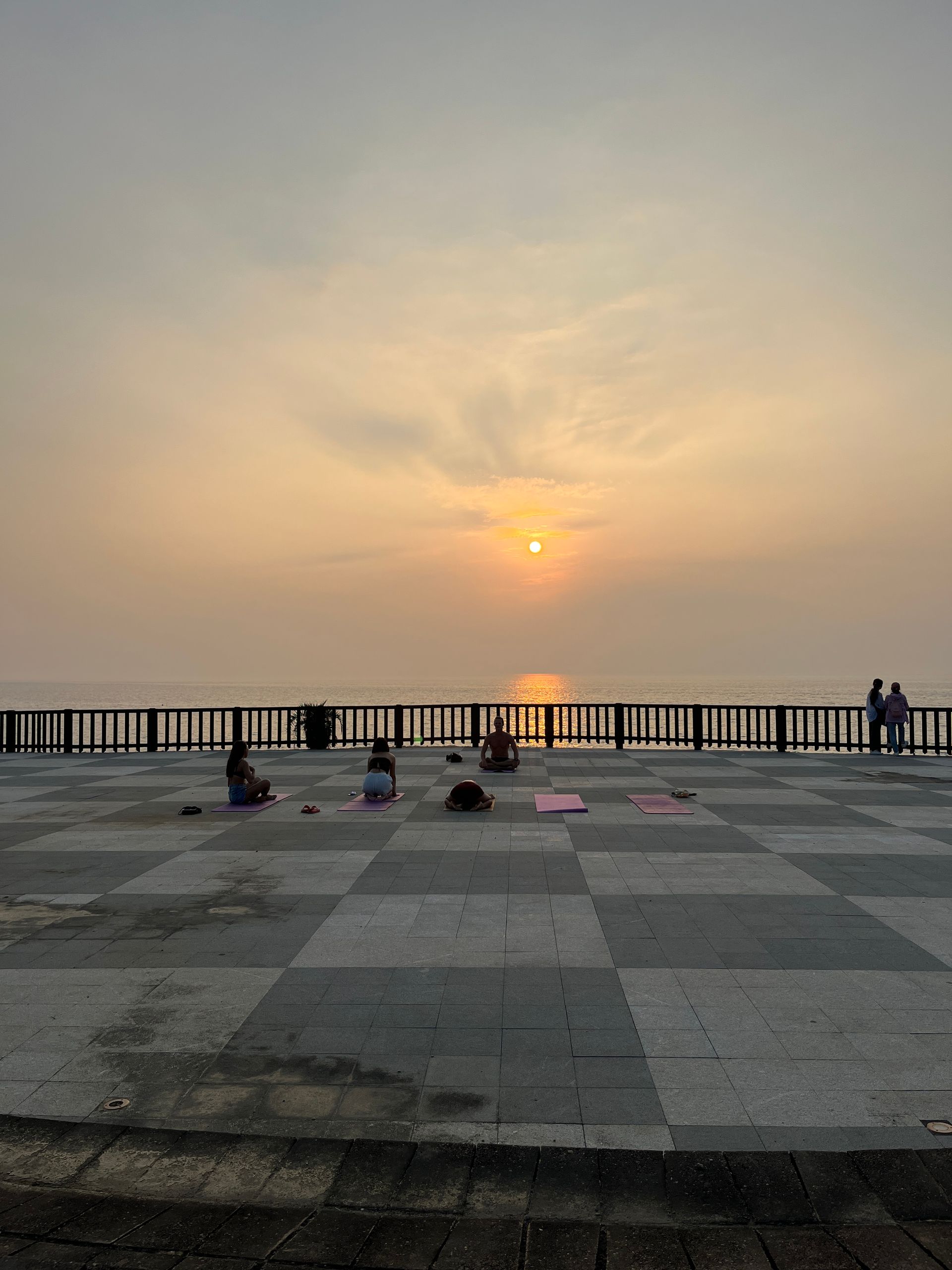 A group of people are sitting on a checkered floor watching the sun set over the ocean.