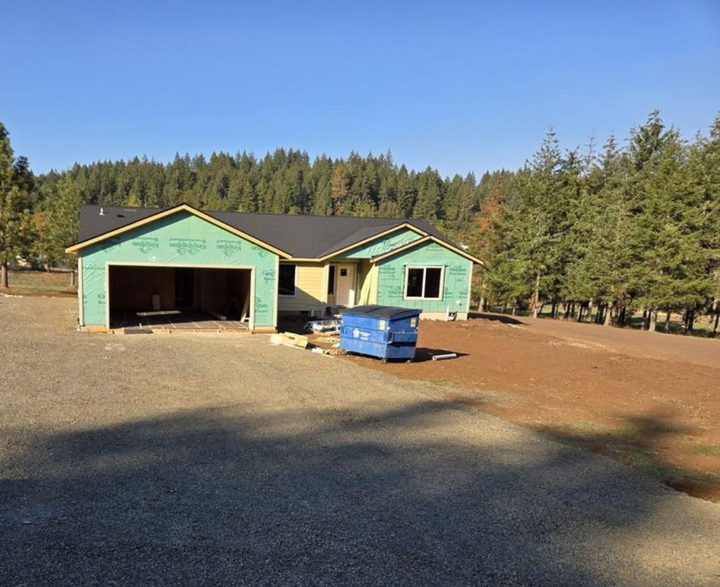 House under construction, green sheathing, open garage, blue dumpster, gravel driveway, trees in background.