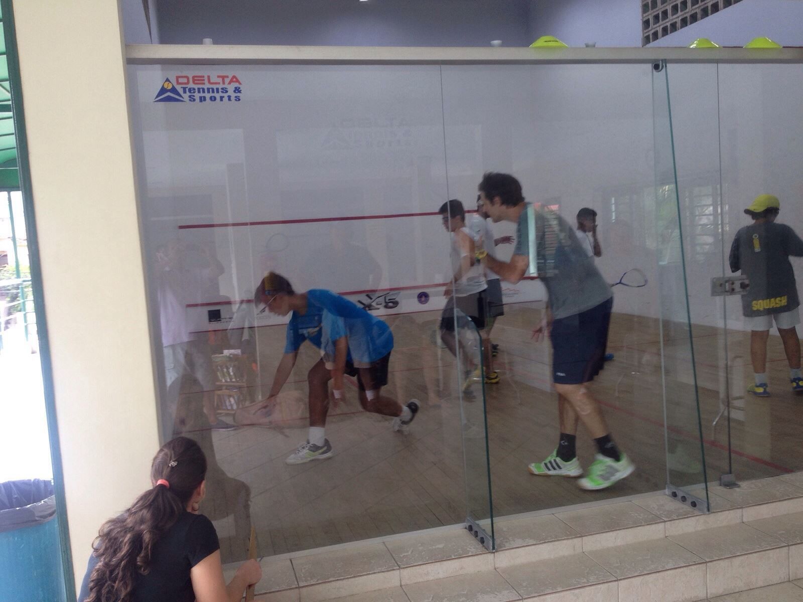 um grupo de pessoas joga squash em uma academia atrás de uma parede de vidro.