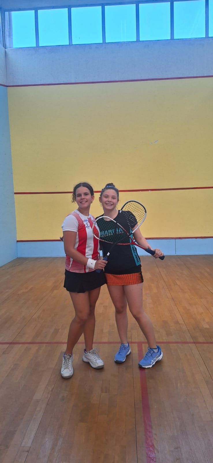 Duas mulheres estão de pé, uma ao lado da outra, em uma quadra de squash, segurando raquetes.