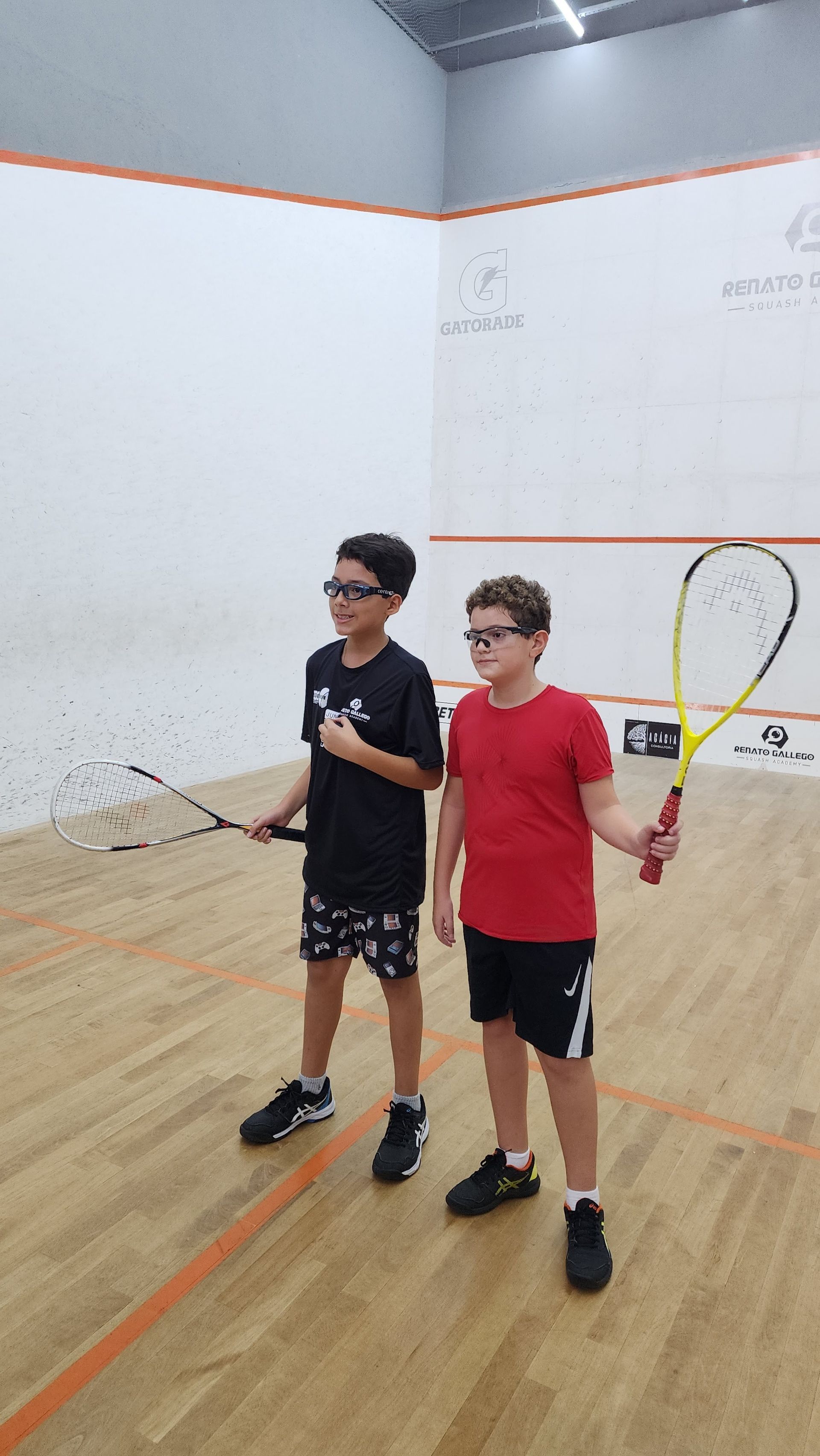 Dois meninos estão em uma quadra de squash segurando raquetes.