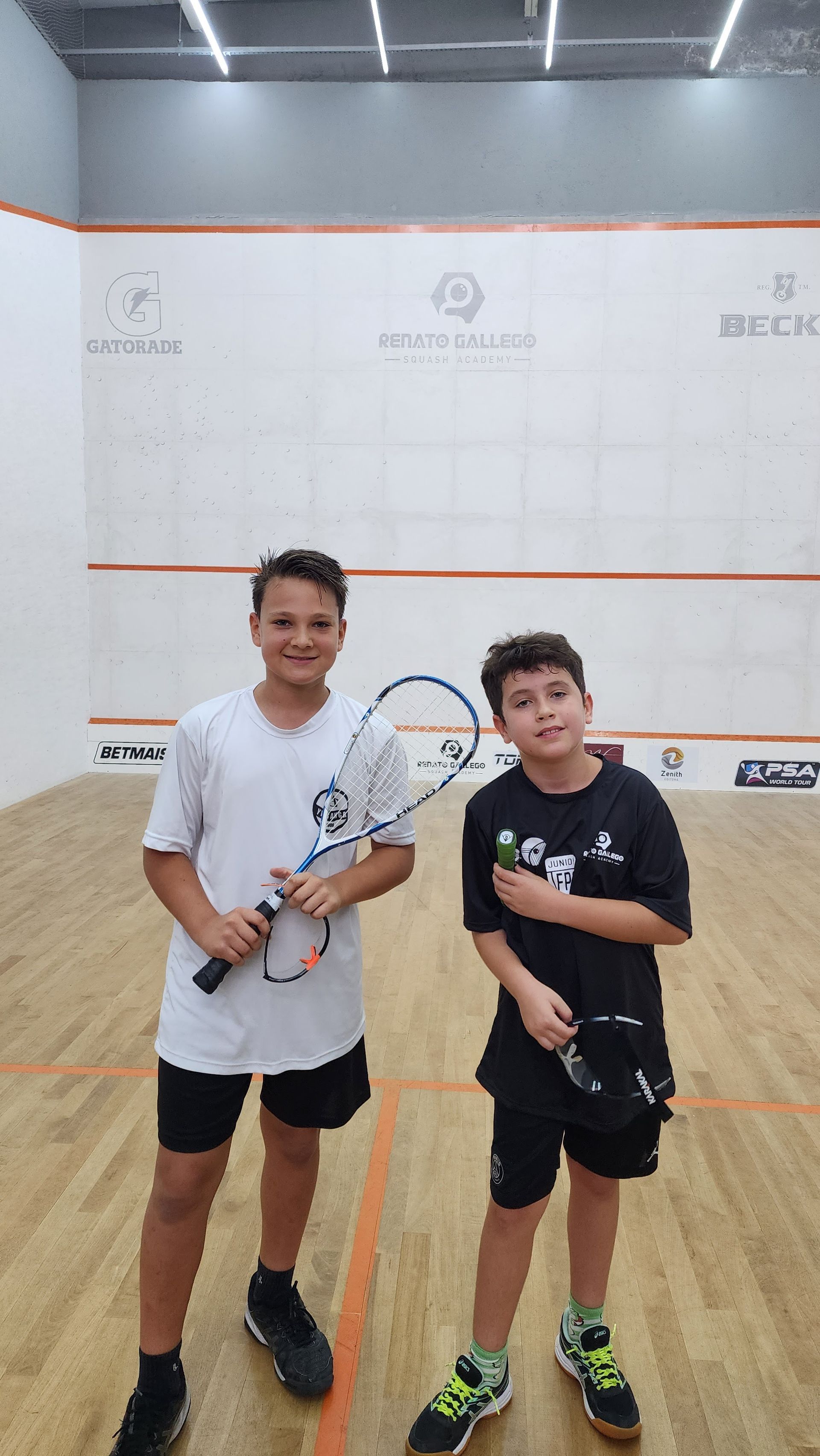 Dois meninos estão em uma quadra de squash segurando raquetes.