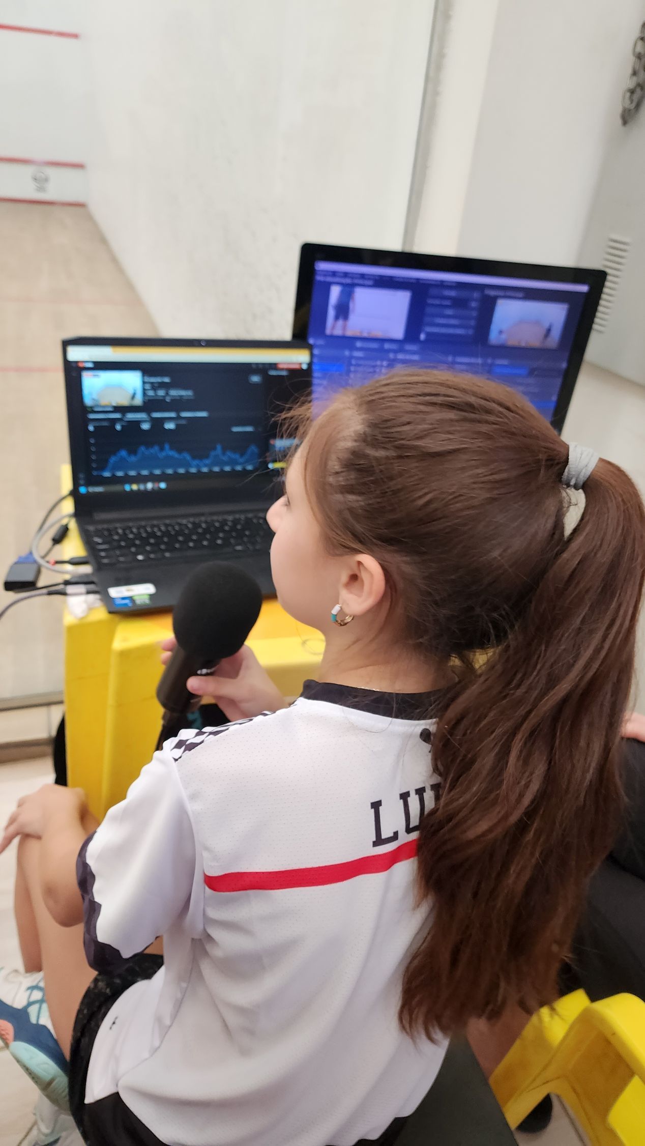 Uma garota está sentada em frente a dois laptops e um microfone.