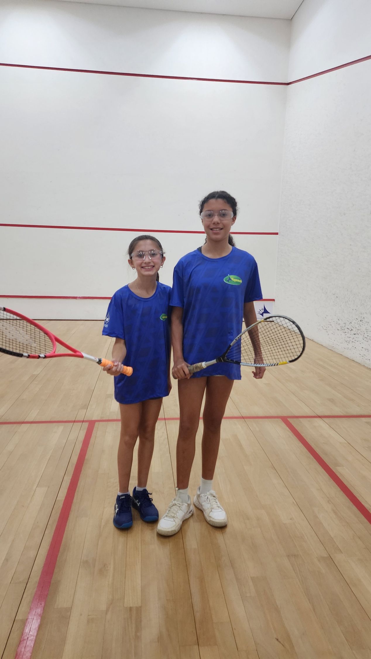 Duas meninas estão em uma quadra de squash segurando raquetes de tênis.