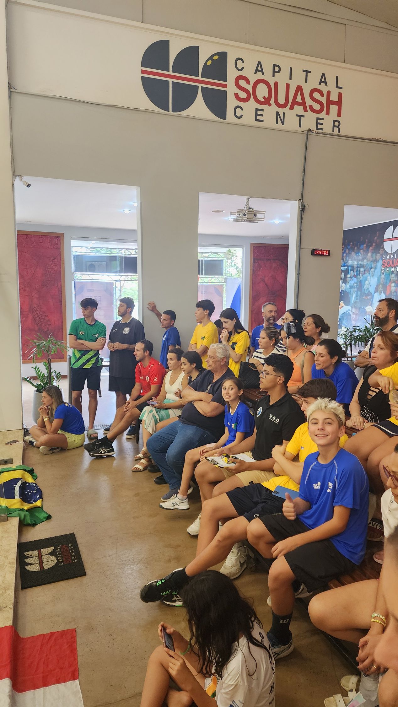 um grupo de pessoas está sentado em uma sala com uma placa que diz centro de squash capital.