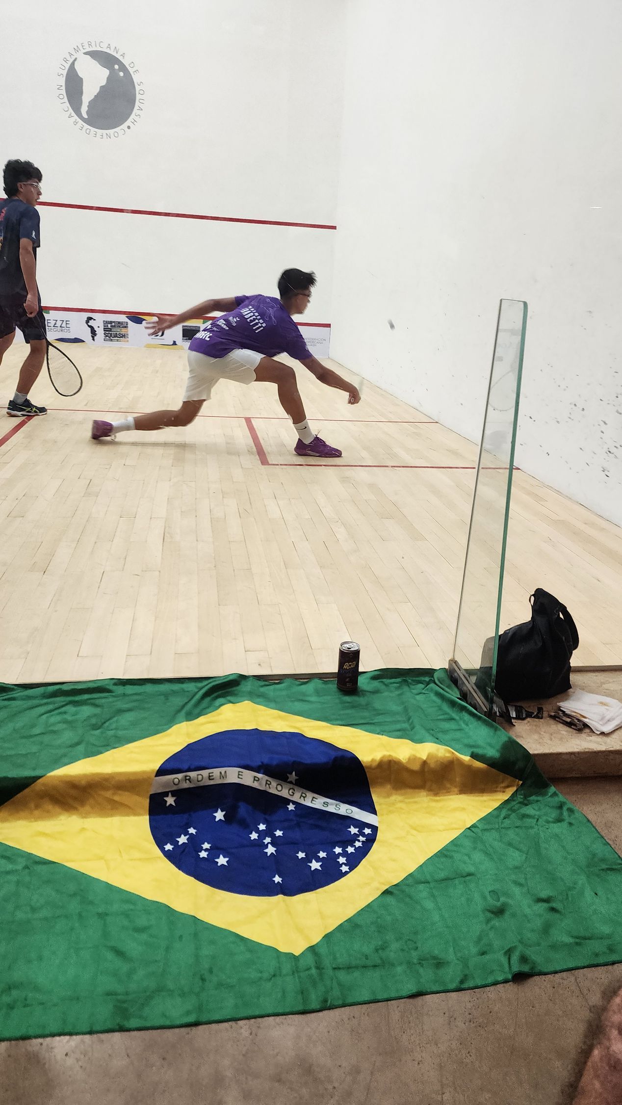 dois homens jogam squash em uma quadra com uma bandeira brasileira em primeiro plano.