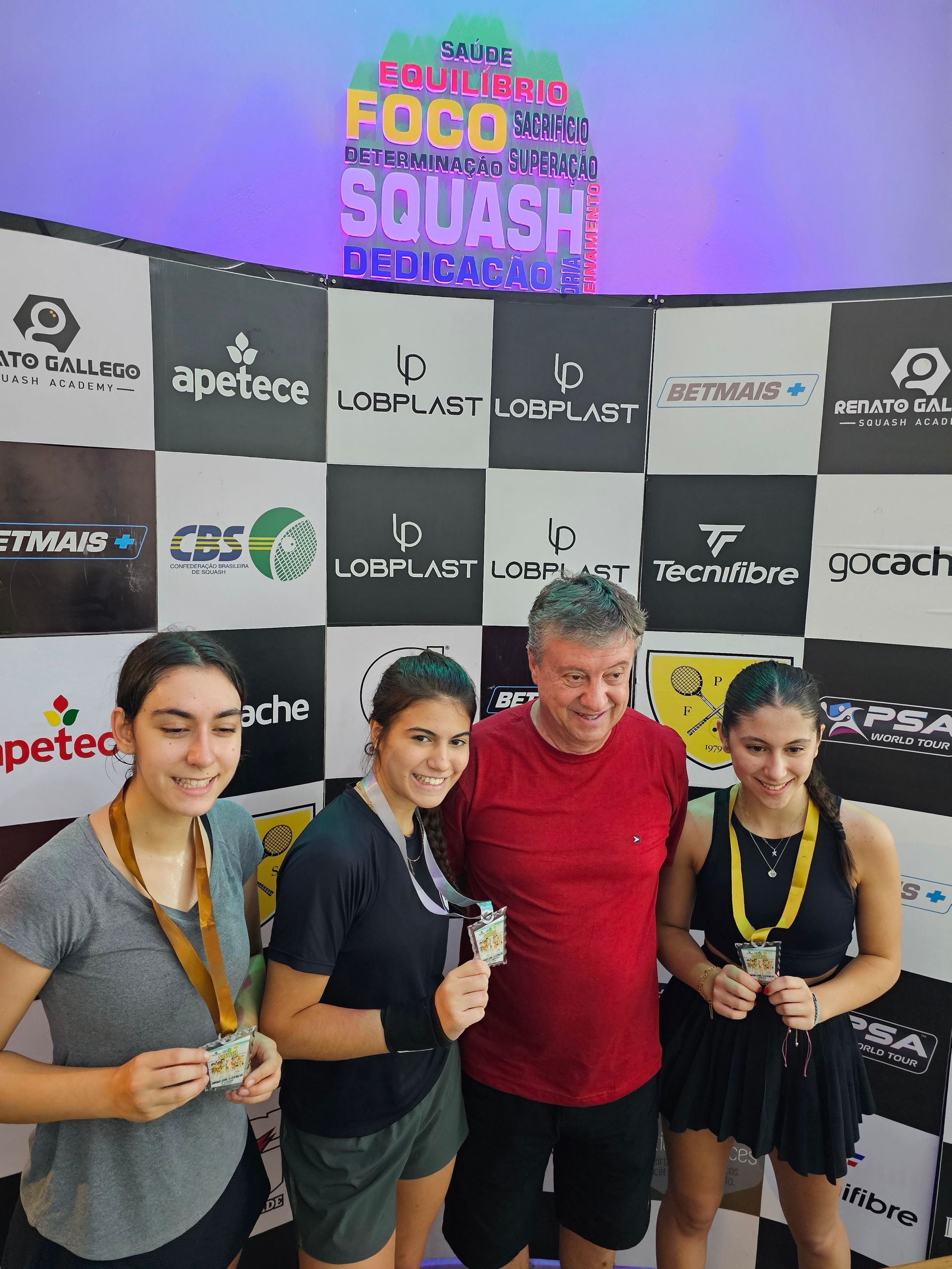 Um grupo de pessoas em pé, uma ao lado da outra, em frente a uma parede, segurando medalhas.