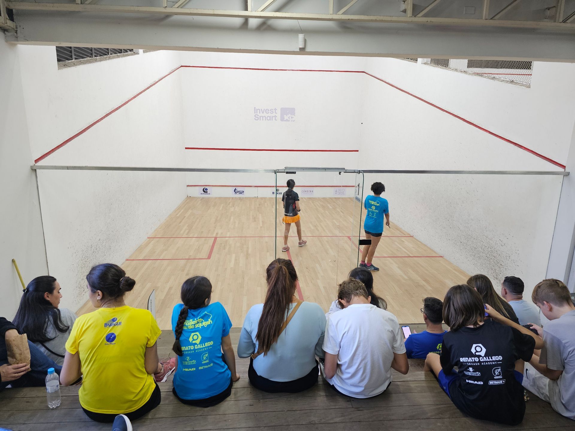 Um grupo de pessoas está sentado no chão assistindo a um jogo de squash