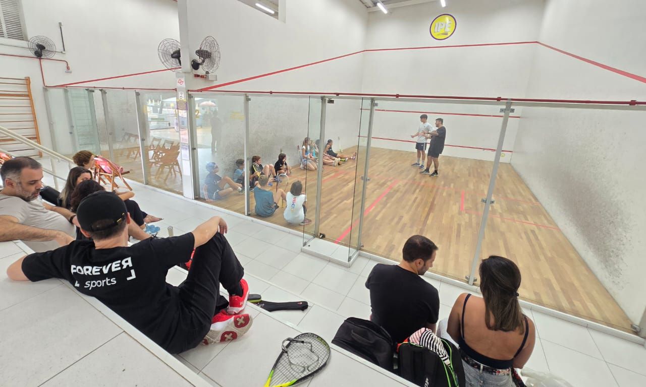 Um grupo de pessoas está sentado na beira de uma quadra de squash.