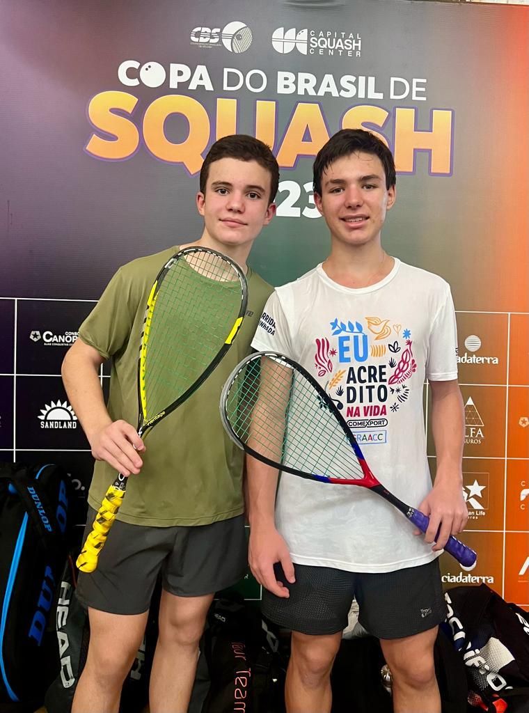 Dois jovens estão de pé, um ao lado do outro, segurando raquetes de squash.
