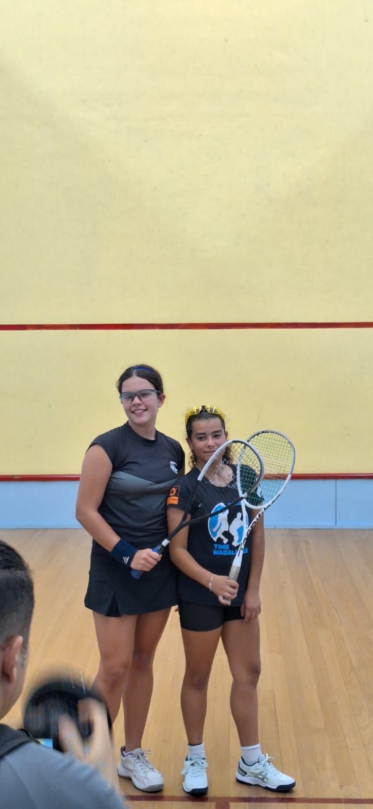 Duas mulheres estão de pé, uma ao lado da outra, em uma quadra de squash, segurando raquetes de tênis.
