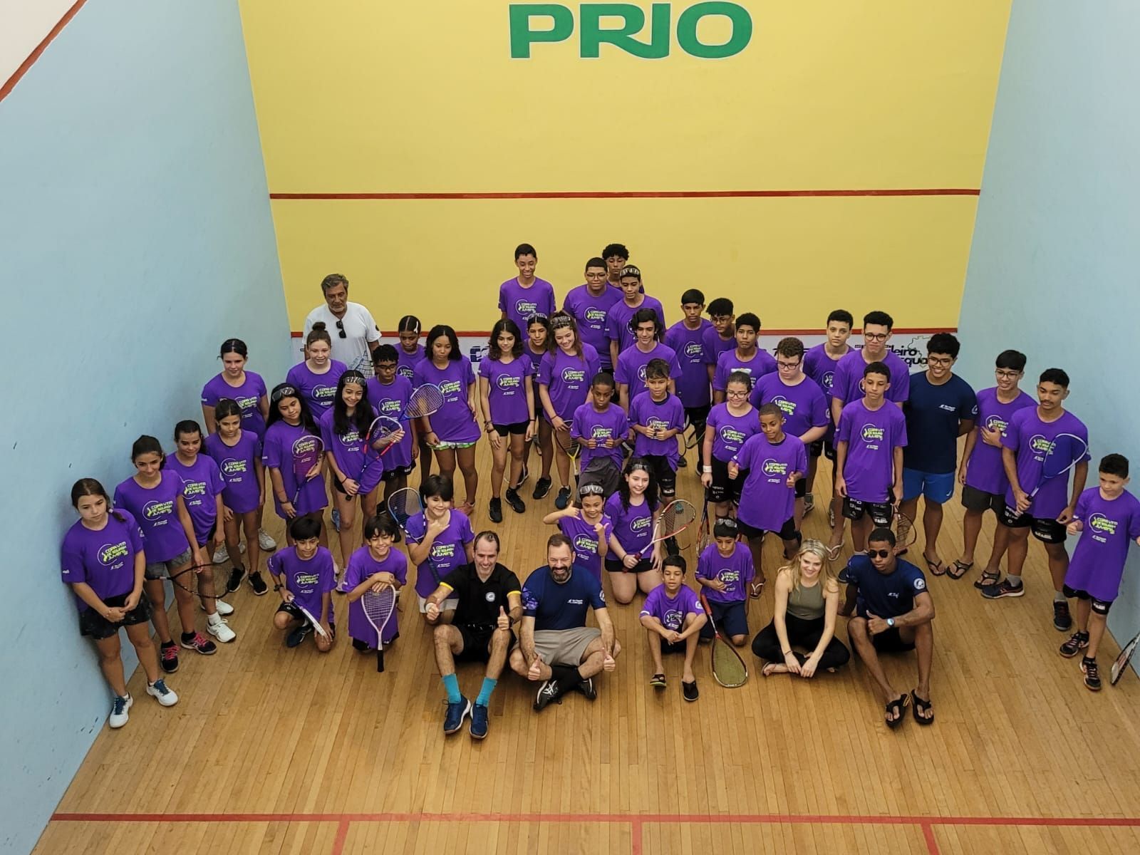 Um grupo de pessoas em uma quadra de squash está posando para uma foto em frente a uma placa que diz prio