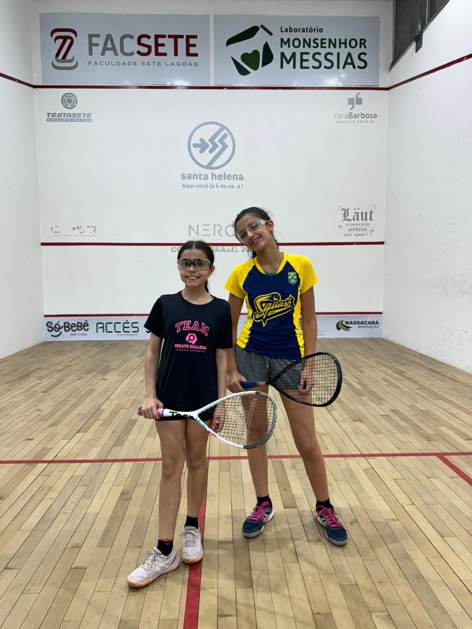 Duas meninas estão em uma quadra de squash segurando raquetes de tênis