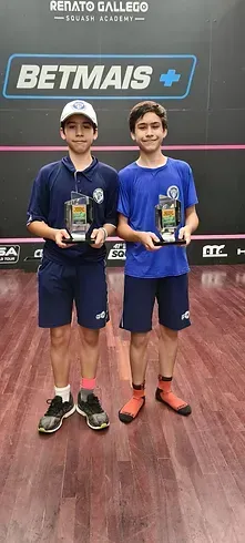 Dois meninos estão de pé, um ao lado do outro, em um piso de madeira, segurando troféus.