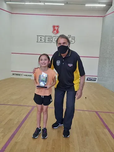 Um homem usando uma máscara está ao lado de uma jovem segurando um troféu em uma quadra de squash.