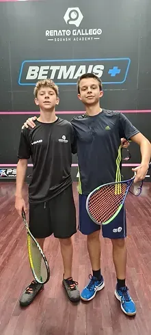 Dois jovens estão de pé, um ao lado do outro, segurando raquetes de tênis.