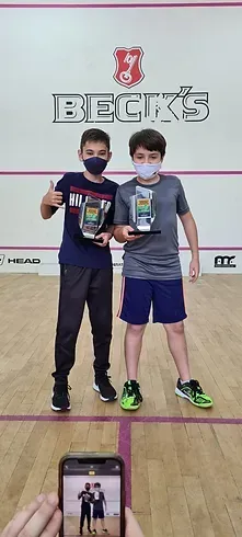 Uma pessoa está tirando uma foto de dois meninos segurando troféus em uma quadra de squash.