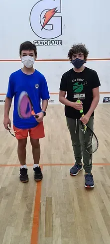 Dois meninos usando máscaras estão segurando raquetes de tênis em uma quadra.