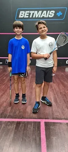 Dois meninos estão em uma quadra de squash segurando raquetes.