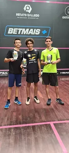Três jovens estão de pé, um ao lado do outro, em uma quadra de squash, segurando troféus.