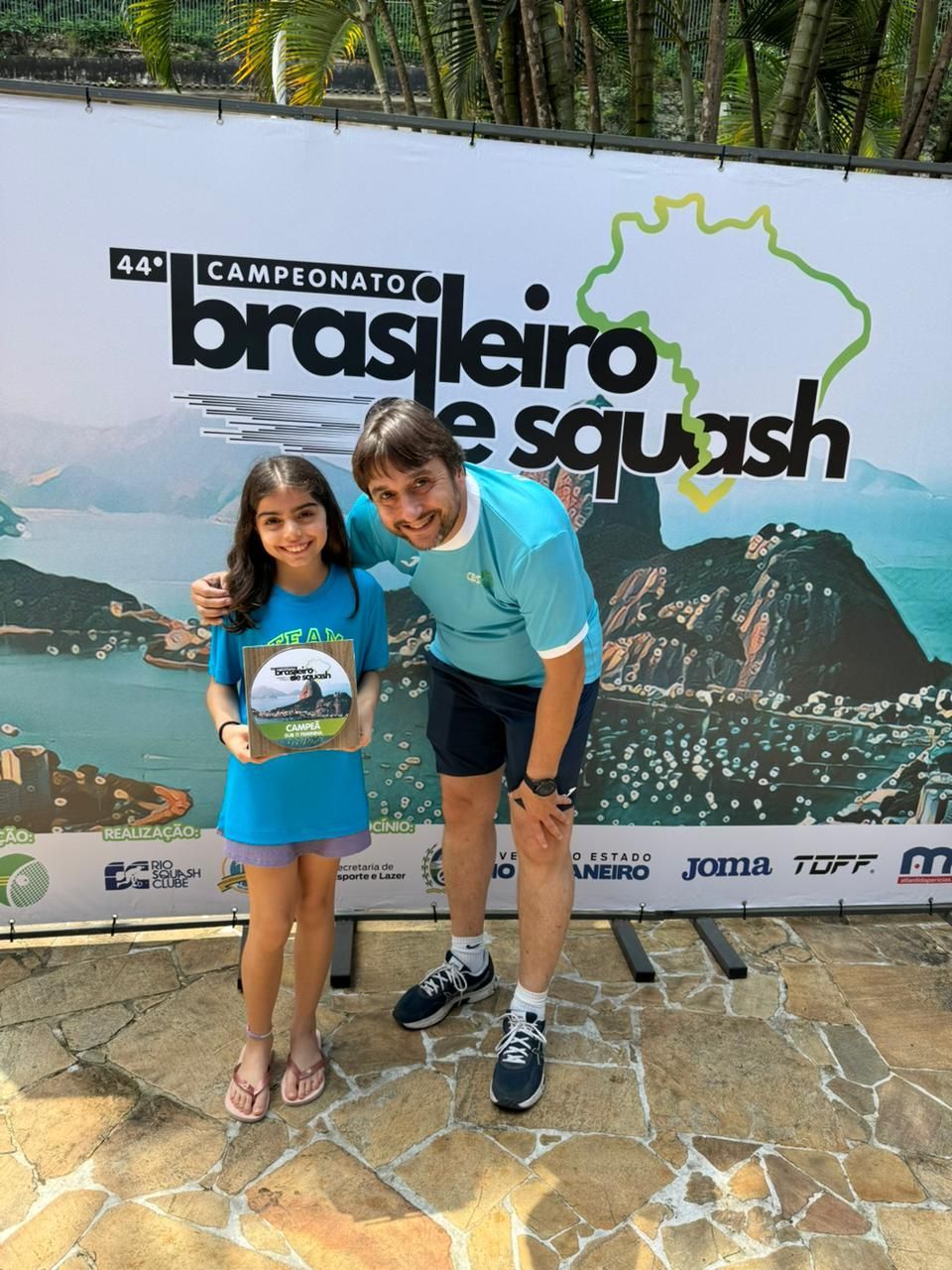 Um homem e uma menina estão posando em frente a uma placa que diz brasileiro e squash