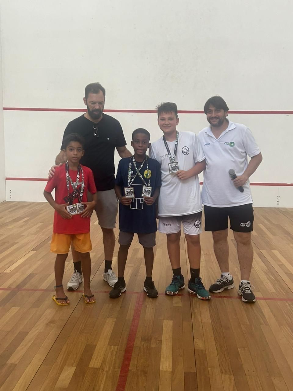 Um grupo de pessoas em pé sobre um piso de madeira segurando medalhas em uma quadra de squash