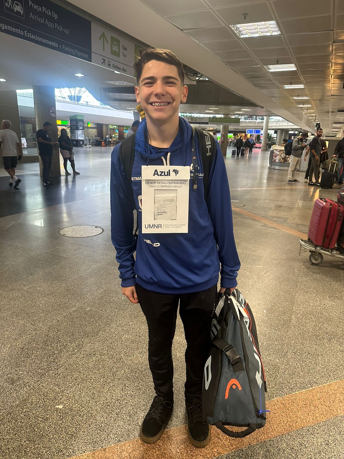Um jovem está parado em um aeroporto com uma mochila e uma bolsa de tênis.
