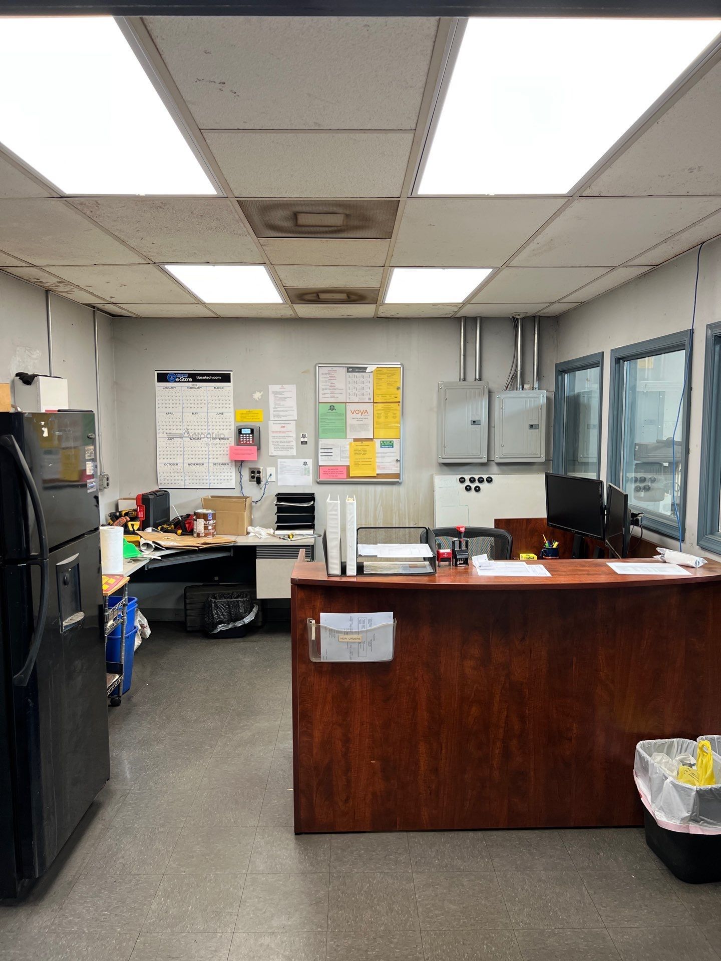 Office interior with desk, fridge, notices, windows, and ceiling lights.