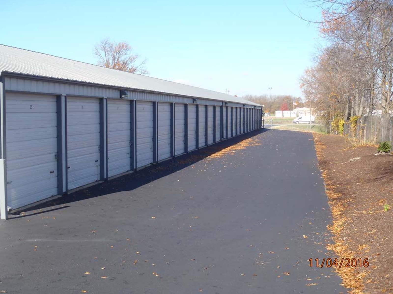 Row of gray storage unit doors along a paved lane beside bare trees and a clear sky