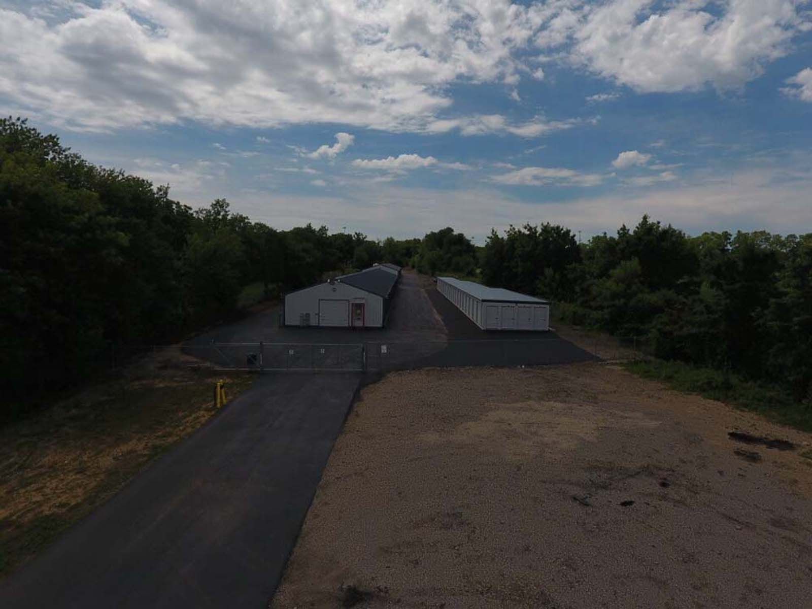 Rural industrial site with two white buildings, paved drive, and trees under a cloudy sky