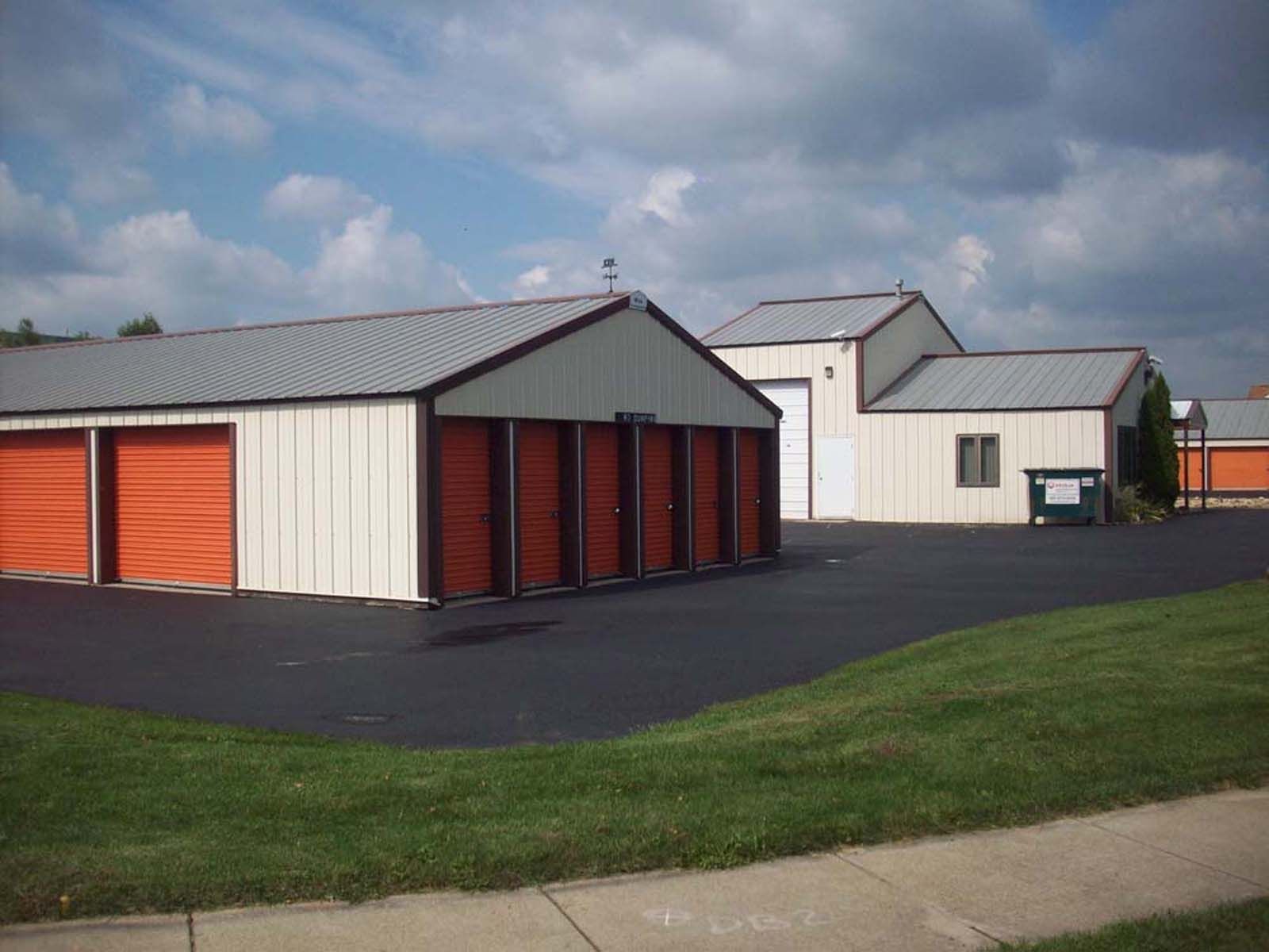 Storage unit buildings with red doors beside a paved lot and grass under a cloudy sky