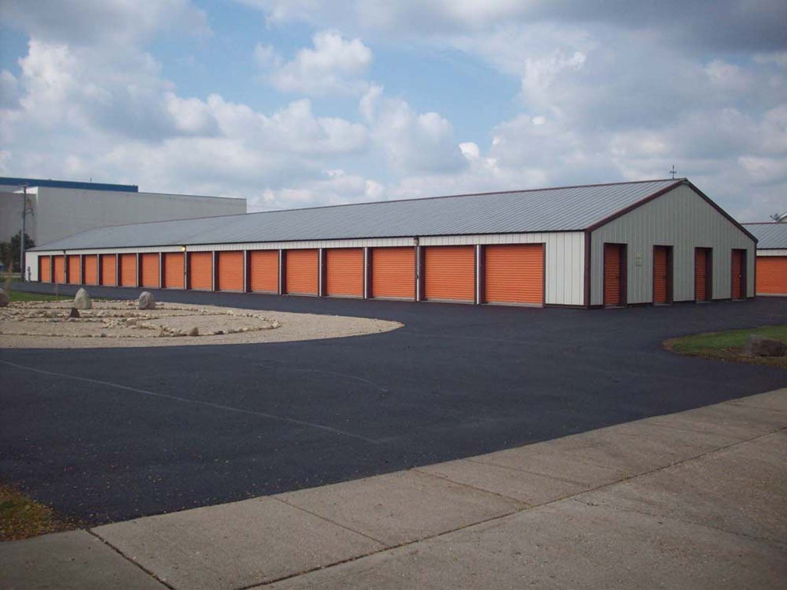 Long storage building with orange roll-up doors and a gray roof under a cloudy sky