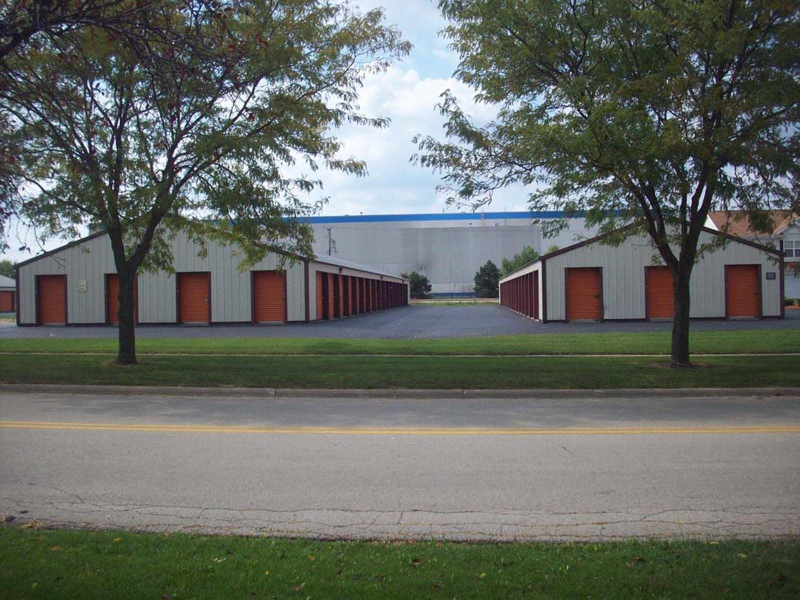 Row of white storage units with red doors behind a grassy roadside, framed by trees