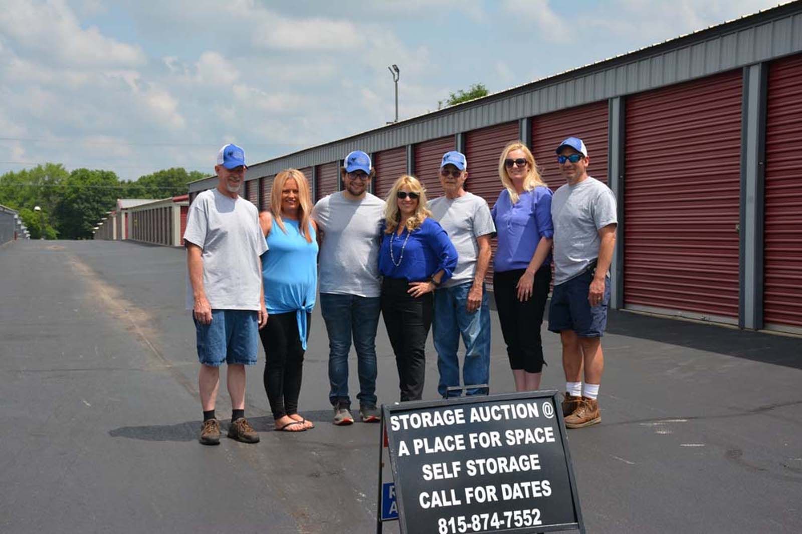 Seven people in hard hats stand by a self-storage facility sign on a sunny paved lot.
