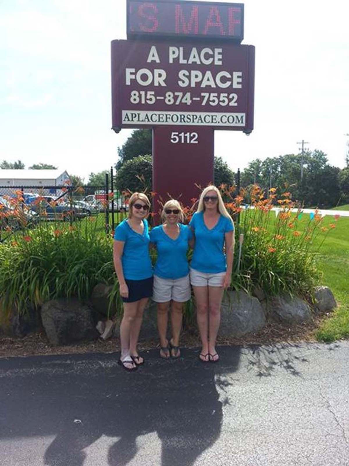Three people in blue shirts posing by a “A Place for Space” sign at a flower-filled roadside stop.