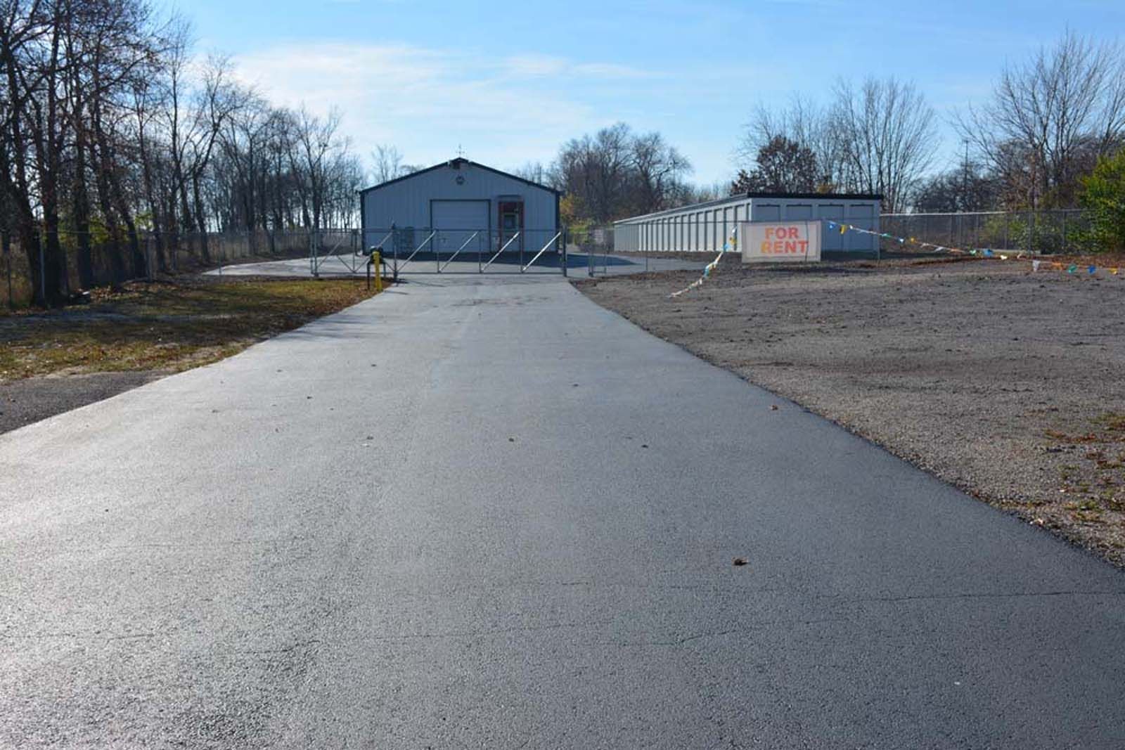 Paved driveway leading to a gray warehouse building with a gated entrance and bare trees nearby
