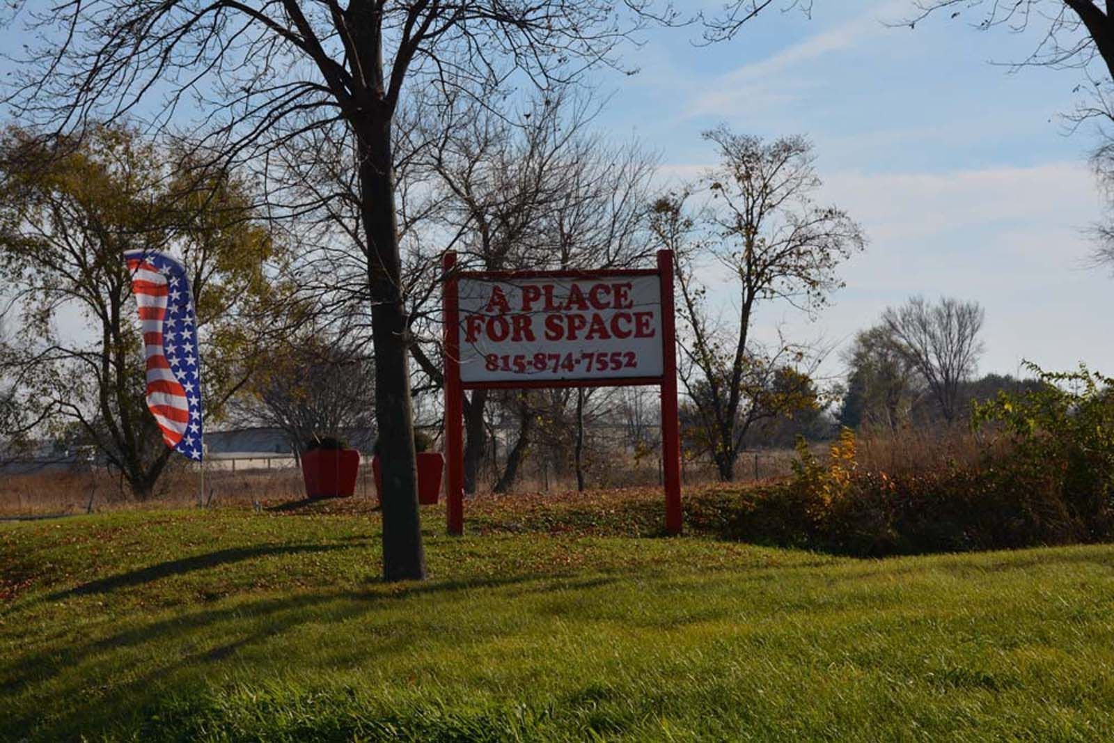 Sign for “A Place for Space” beside a grassy roadside with trees and an American flag banner