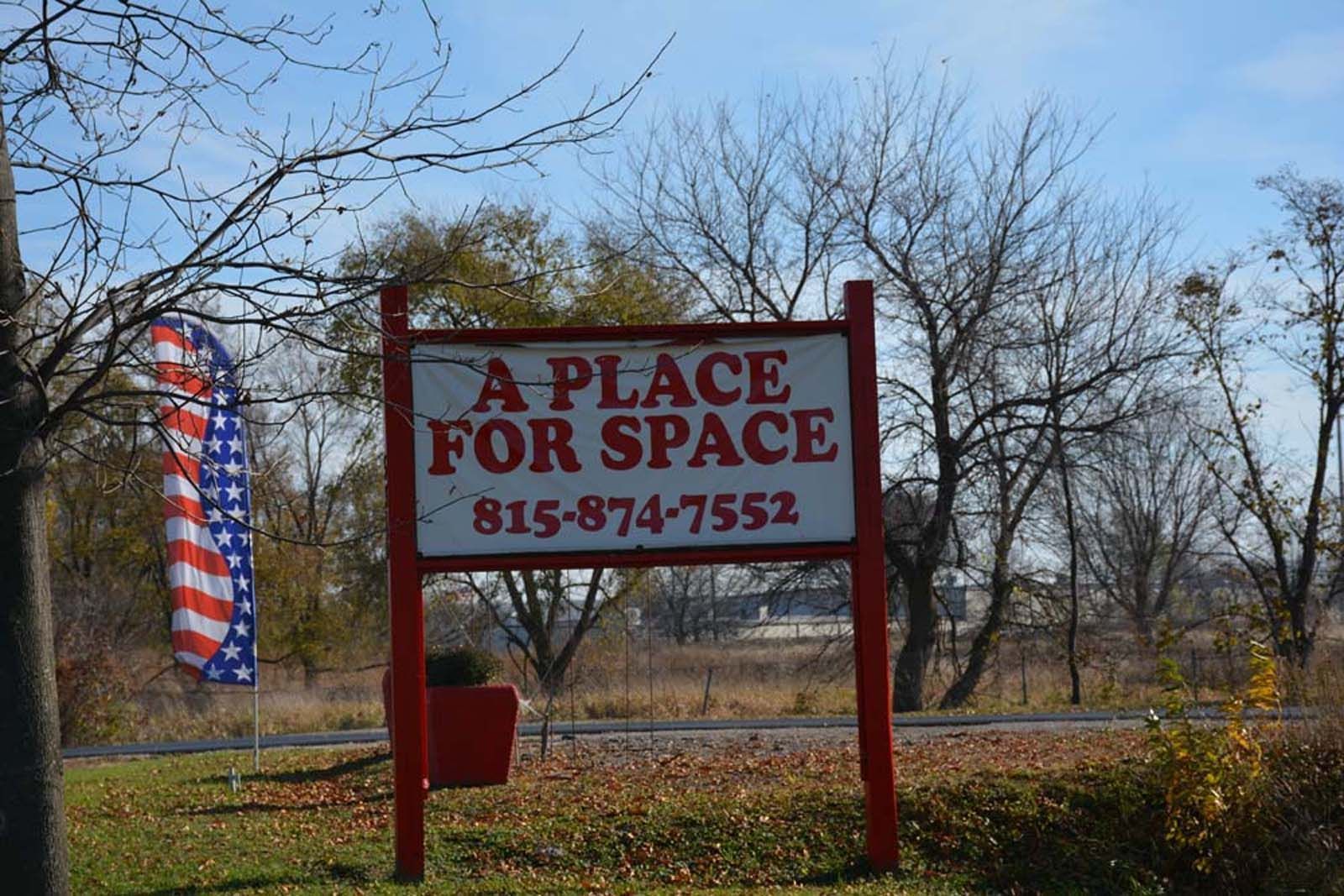 Red sign reading “A Place for Space” with phone number, beside an American flag in a grassy roadside area.