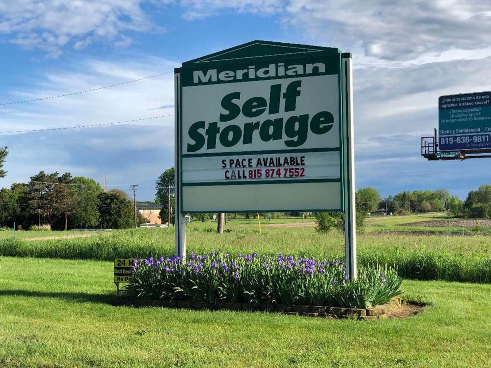 Meridian Self Storage roadside sign in a grassy field with purple flowers under a cloudy sky