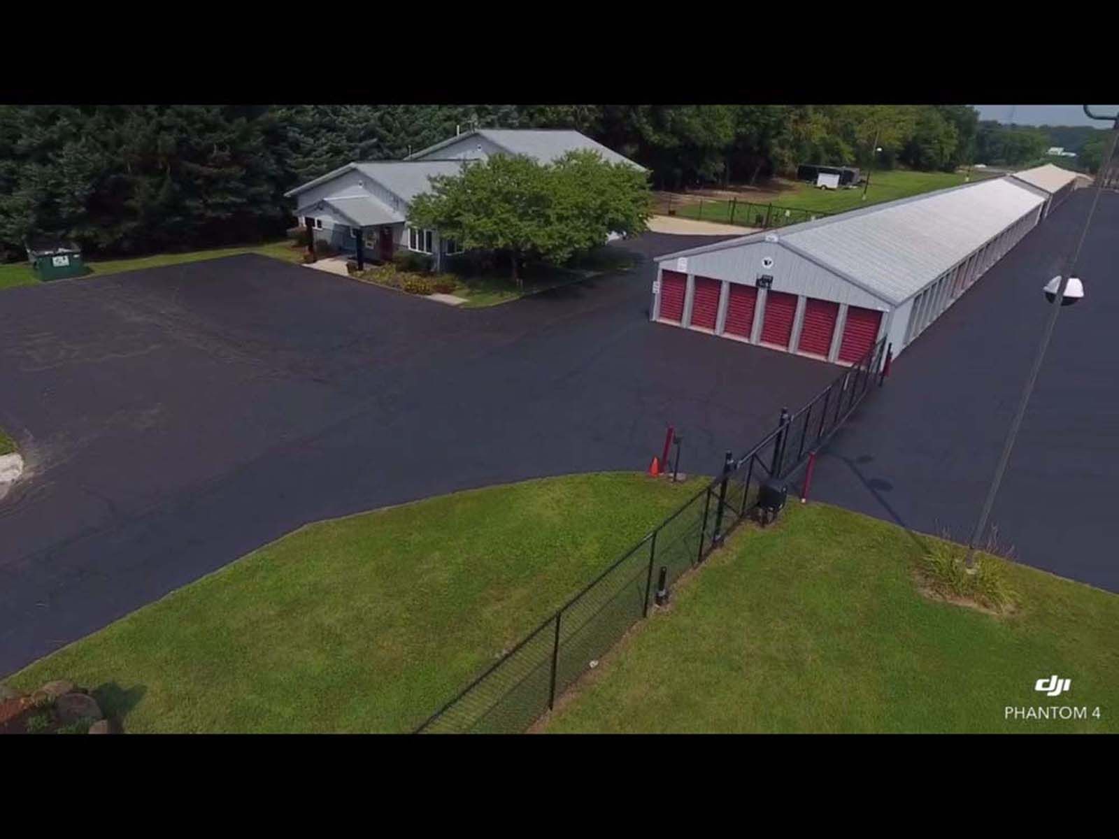 Aerial view of a storage facility with white buildings, red doors, black pavement, and a fenced grassy area.