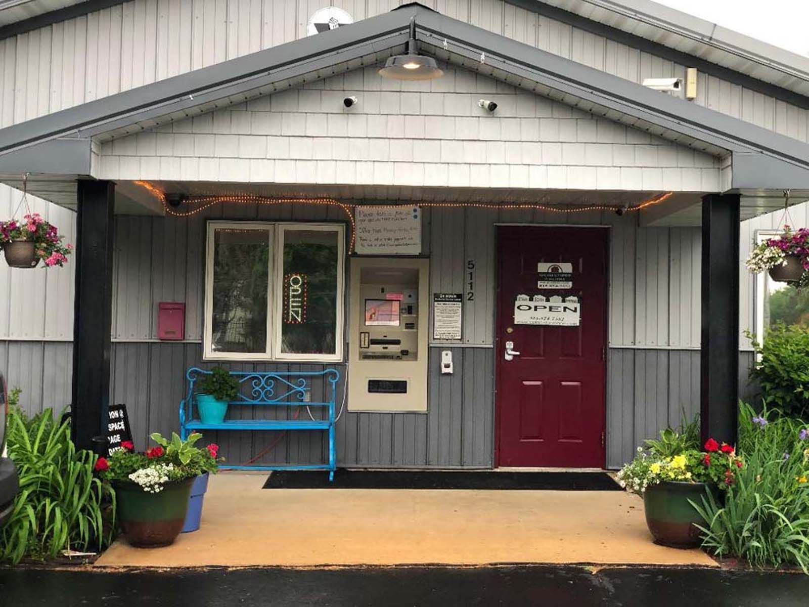 Small gray storefront with red door, blue bench, and flower planters under a gabled roof