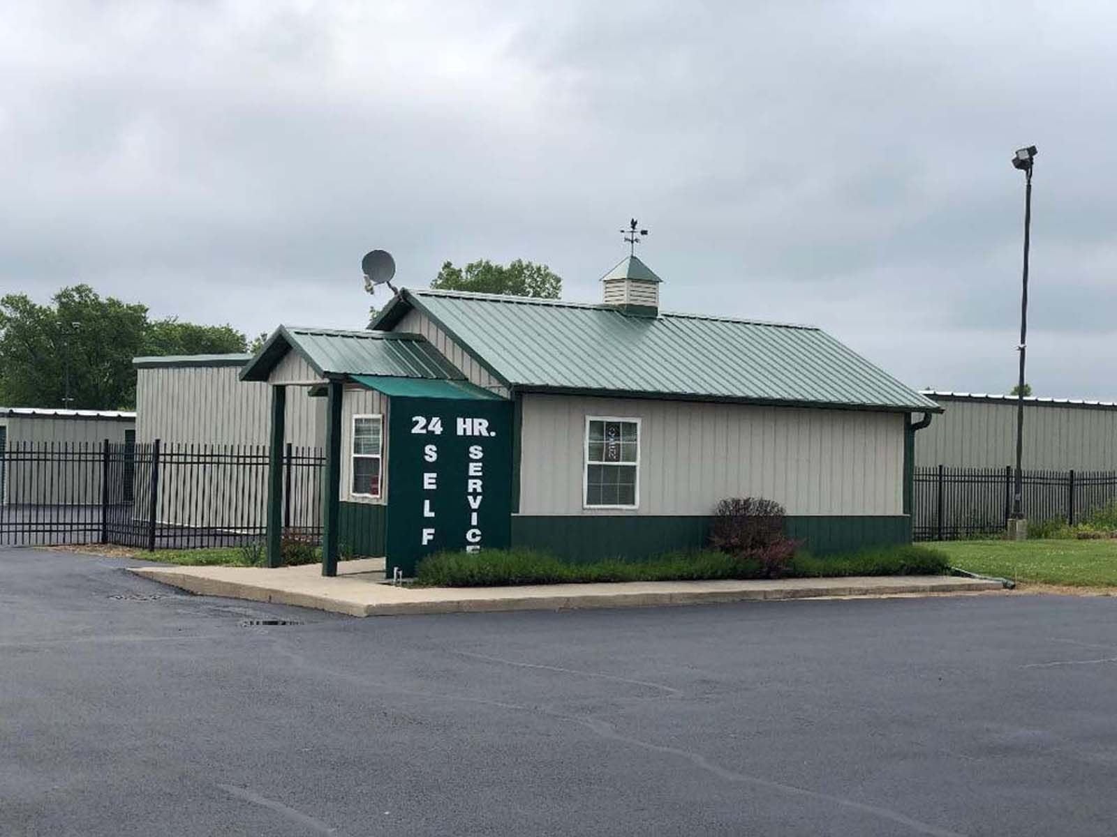 Small green guard shack with a green roof in a fenced parking lot, next to a paved road.