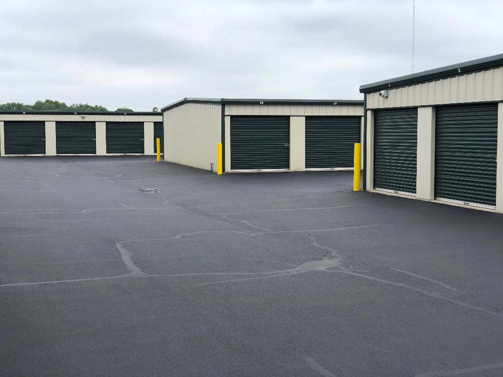 Storage unit row with closed green doors and yellow bollards on an empty paved lot under a cloudy sky