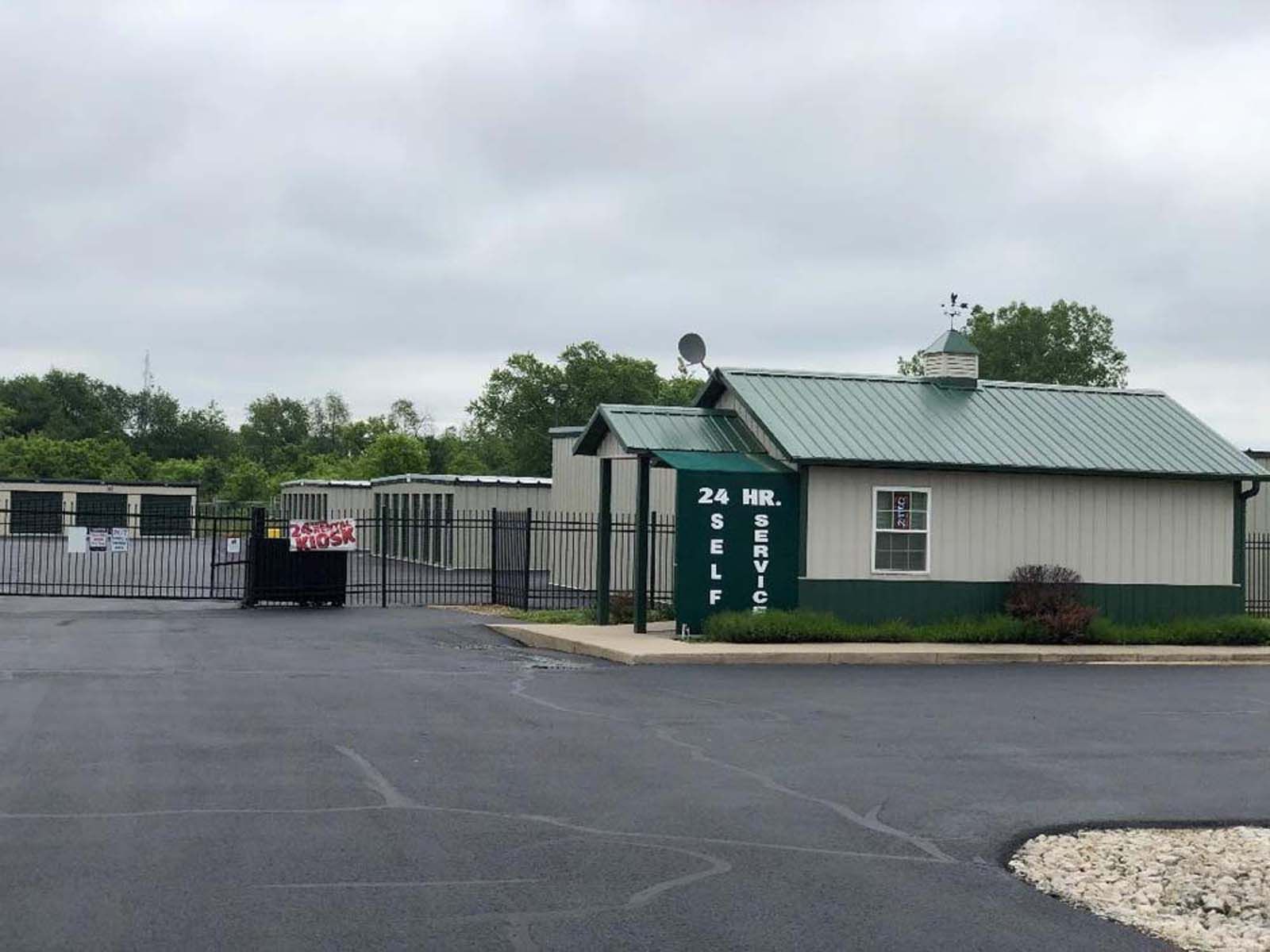 Small green building with fence and trash bins beside an empty asphalt lot under cloudy sky