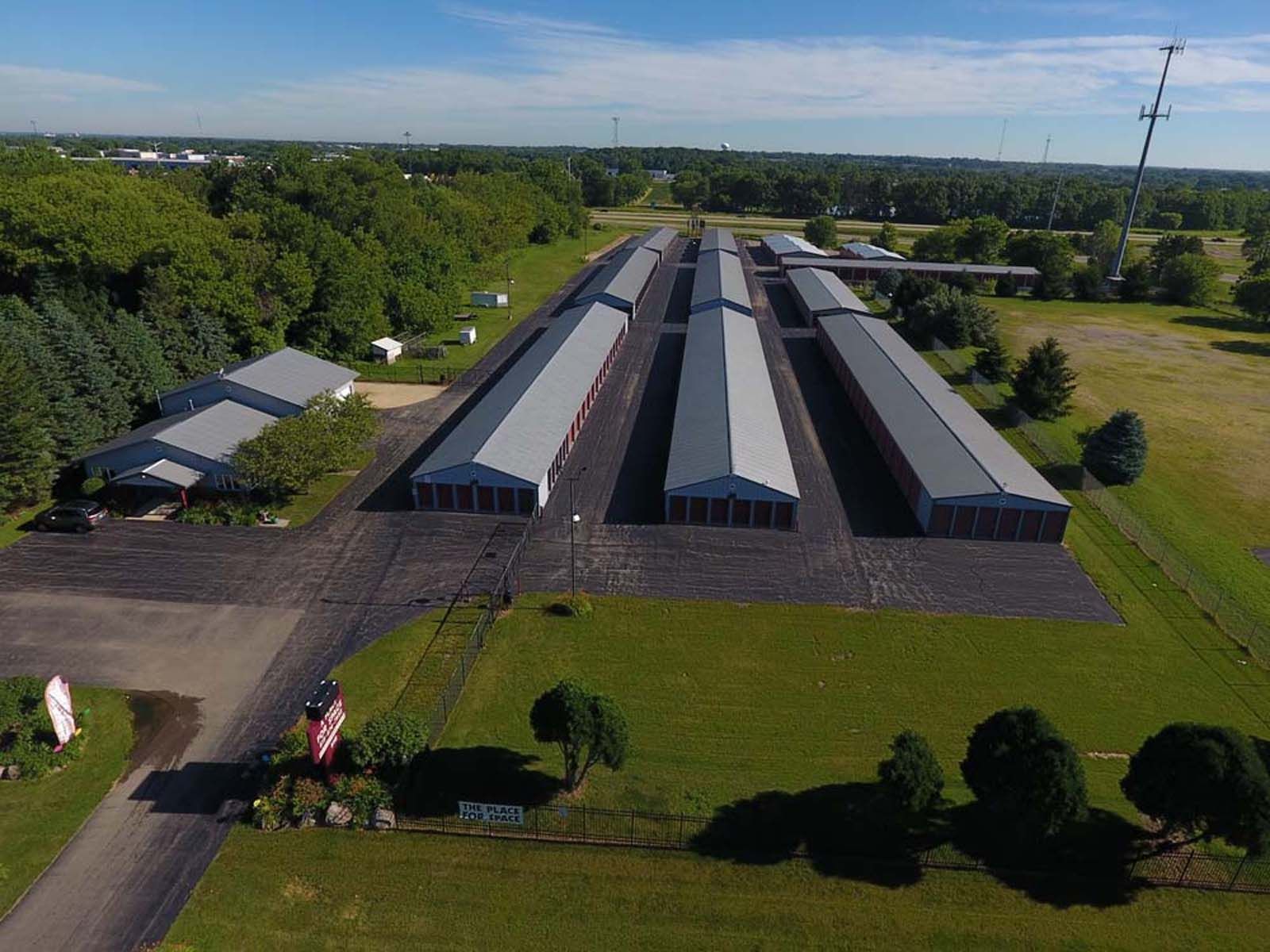 Aerial view of several long gray-roofed buildings beside trees, grass, and a paved road.